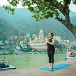 Woman practicing yoga by a serene riverside with statues in the backdrop, Rishikesh, India.