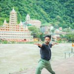 A man performing yoga on a riverbank in Rishikesh with iconic temples in the background.